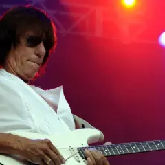 Jeff Beck, con occhiali da sole e una maglietta bianca, suona la chitarra elettrica con il suo inconfondibile stile. La sua musica, un'eredità del **the Jeff Beck group**, risuona sotto le luci rosse del palco.