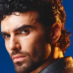 Volto intenso di un uomo con capelli ricci scuri e barba, illuminato da una luce dorata. Ricorda i cantanti di una volta.