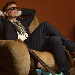Gianni Rojatti, con occhiali da sole e un completo elegante, si rilassa su una poltrona con una chitarra ai piedi.