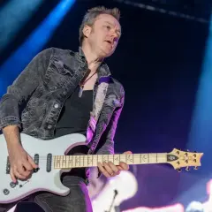 Chitarrista dei Deep Purple, con giacca di jeans scuro e chitarra bianca, sul palco illuminato da luci blu.