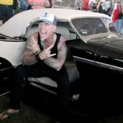 Metallica James Hetfield, con cappellino e canotta, fa il segno delle corna seduto in un'auto d'epoca nera.