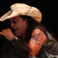 Pino Scotto, icona rock degli anni '80, canta con passione indossando un cappello da cowboy e occhiali da sole.