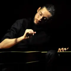 Giovanni Baglioni, con la sua chitarra, crea un'atmosfera intima e intensa. La sua mano destra accarezza le corde, mentre il volto è immerso nella penombra.