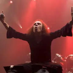 Ronnie James Dio, icona heavy metal, sul palco con le braccia alzate, microfono in mano e il segno delle corna. La sua voce potente risuona, sfidando la morte.
