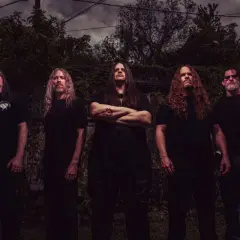 I membri dei Cannibal Corpse posano in un ambiente esterno cupo, con alberi e cielo nuvoloso sullo sfondo.