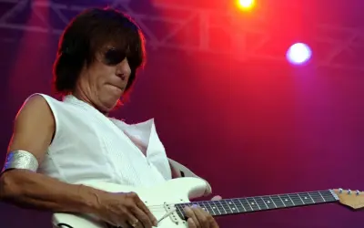 Jeff Beck, con occhiali da sole e una maglietta bianca, suona la chitarra elettrica con il suo inconfondibile stile. La sua musica, un'eredità del **the Jeff Beck group**, risuona sotto le luci rosse del palco.