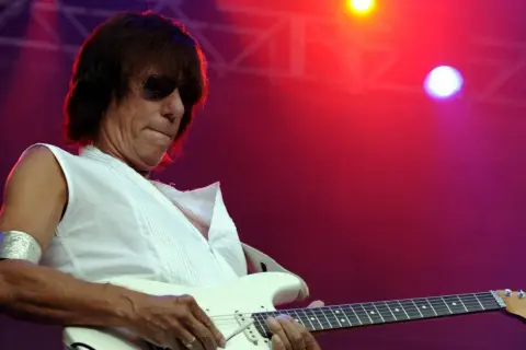 Jeff Beck, con occhiali da sole e una maglietta bianca, suona la chitarra elettrica con il suo inconfondibile stile. La sua musica, un'eredità del **the Jeff Beck group**, risuona sotto le luci rosse del palco.