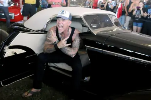 Metallica James Hetfield, con cappellino e canotta, fa il segno delle corna seduto in un'auto d'epoca nera.