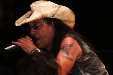 Pino Scotto, icona rock degli anni '80, canta con passione indossando un cappello da cowboy e occhiali da sole.