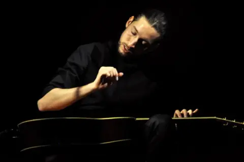 Giovanni Baglioni, con la sua chitarra, crea un'atmosfera intima e intensa. La sua mano destra accarezza le corde, mentre il volto è immerso nella penombra.