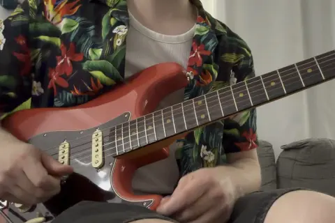 Un chitarrista con una camicia floreale suona una chitarra rossa, ricordando lo stile di Guthrie Govan.