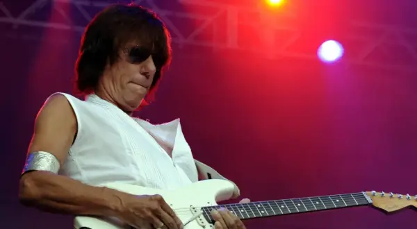 Jeff Beck, con occhiali da sole e una maglietta bianca, suona la chitarra elettrica con il suo inconfondibile stile. La sua musica, un'eredità del **the Jeff Beck group**, risuona sotto le luci rosse del palco.
