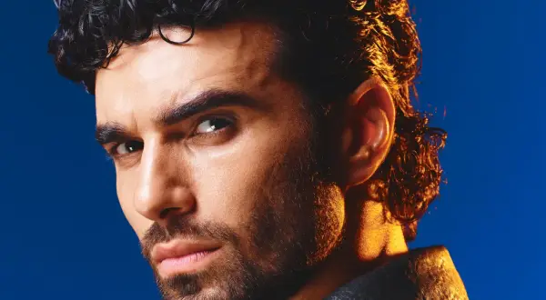 Volto intenso di un uomo con capelli ricci scuri e barba, illuminato da una luce dorata. Ricorda i cantanti di una volta.