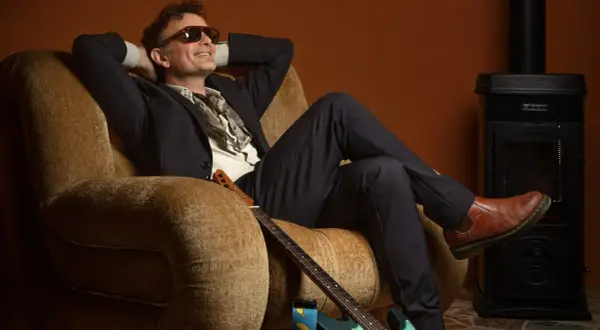 Gianni Rojatti, con occhiali da sole e un completo elegante, si rilassa su una poltrona con una chitarra ai piedi.