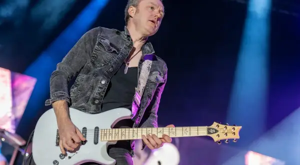 Chitarrista dei Deep Purple, con giacca di jeans scuro e chitarra bianca, sul palco illuminato da luci blu.
