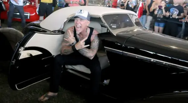 Metallica James Hetfield, con cappellino e canotta, fa il segno delle corna seduto in un'auto d'epoca nera.