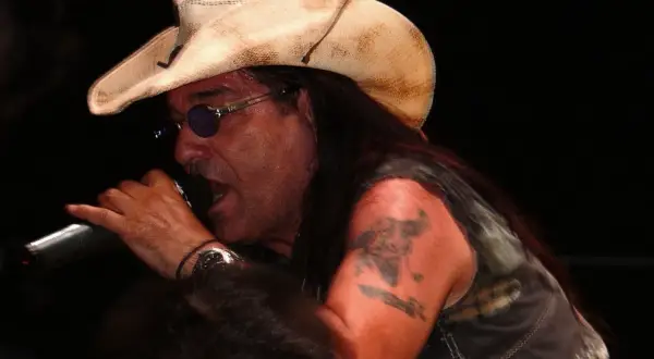 Pino Scotto, icona rock degli anni '80, canta con passione indossando un cappello da cowboy e occhiali da sole.