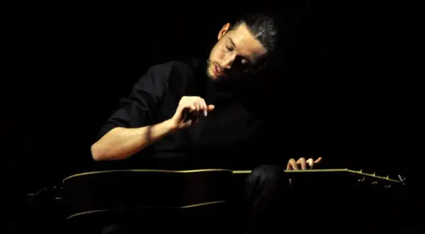 Giovanni Baglioni, con la sua chitarra, crea un'atmosfera intima e intensa. La sua mano destra accarezza le corde, mentre il volto è immerso nella penombra.
