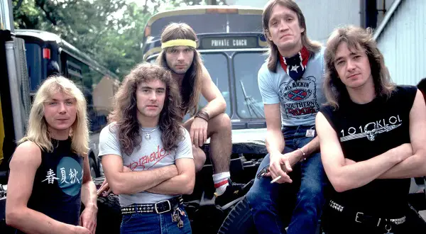 Cinque rocker con capelli lunghi e abbigliamento tipico dei gruppi metal anni 90, posano davanti a un bus privato.