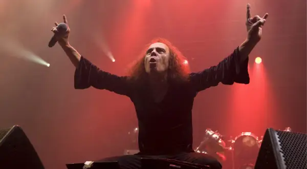 Ronnie James Dio, icona heavy metal, sul palco con le braccia alzate, microfono in mano e il segno delle corna. La sua voce potente risuona, sfidando la morte.