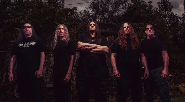 I membri dei Cannibal Corpse posano in un ambiente esterno cupo, con alberi e cielo nuvoloso sullo sfondo.