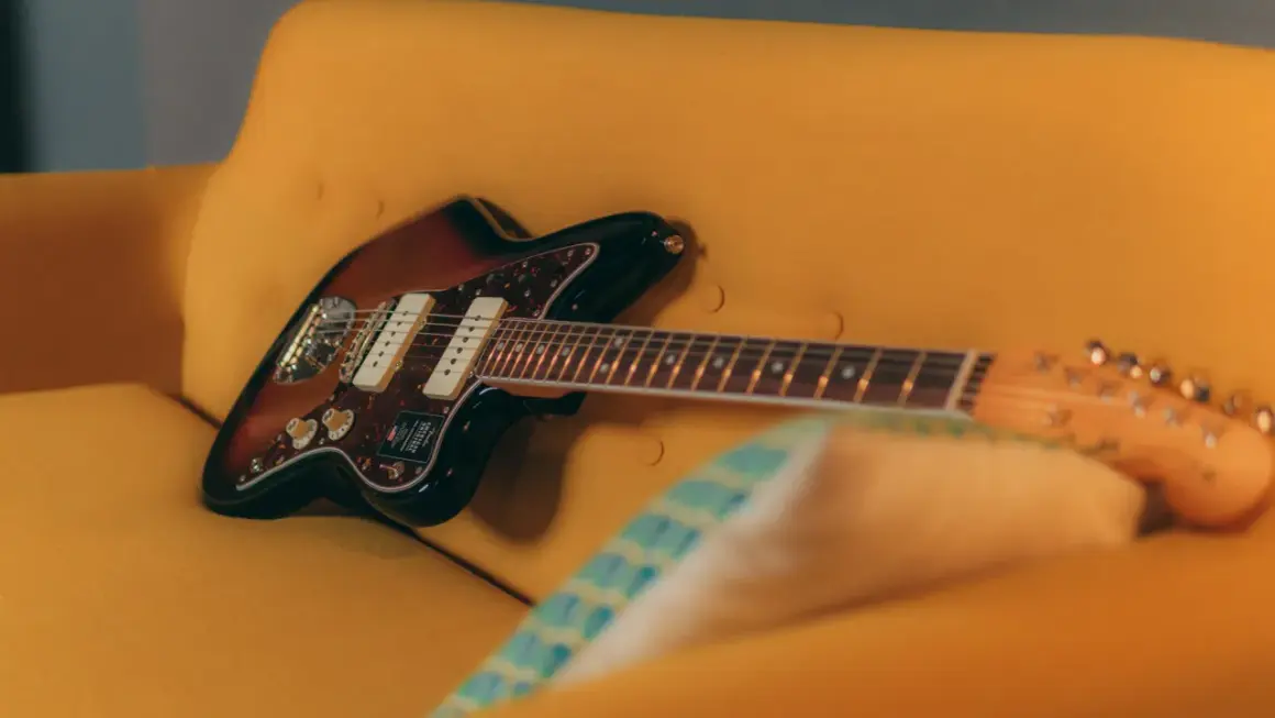 Chitarra Fender Jazzmaster color sunburst appoggiata su un divano giallo. Un duello tra jaguar e jazzmaster, quale suono preferisci?