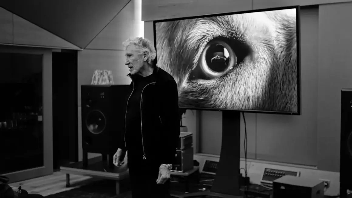 Roger Waters, con un'espressione divertita, osserva un primo piano di un occhio di lupo su uno schermo, forse ispirato da "amused to death".
