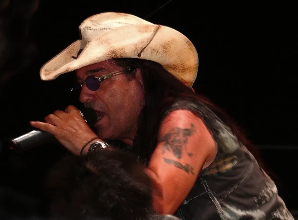 Pino Scotto, icona rock degli anni '80, canta con passione indossando un cappello da cowboy e occhiali da sole.
