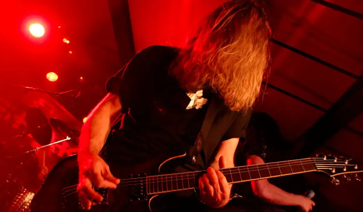 Chitarrista con lunghi capelli biondi, immerso nella luce rossa, crea la base ritmica di un brano con la sua chitarra elettrica.