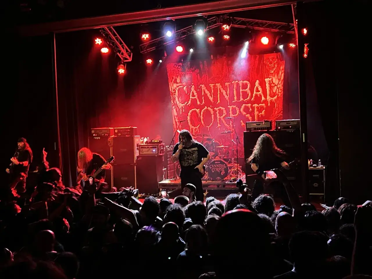 Membri di Cannibal Corpse sul palco, illuminati da luci rosse, davanti a una folla scatenata.