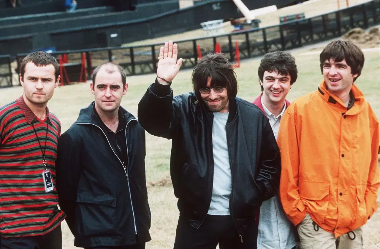I cinque membri degli Oasis, icona degli anni '90, posano sorridenti. Liam saluta con la mano.