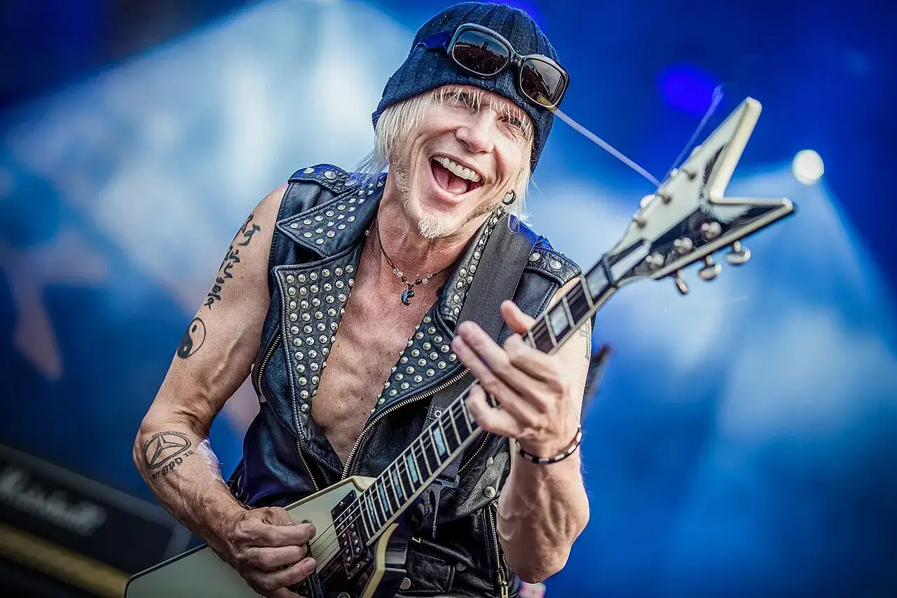 Michael Schenker Group: Michael Schenker sorride mentre suona la sua chitarra a V, con un gilet borchiato e un cappello.