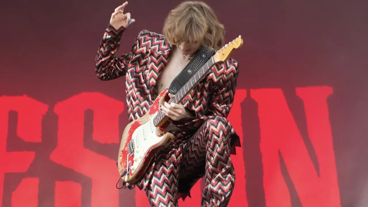 Thomas Raggi, chitarrista dei M&aring;neskin, con la sua chitarra rossa e bianca, indossa una giacca e pantaloni a zig-zag scintillanti su uno sfondo rosso acceso.