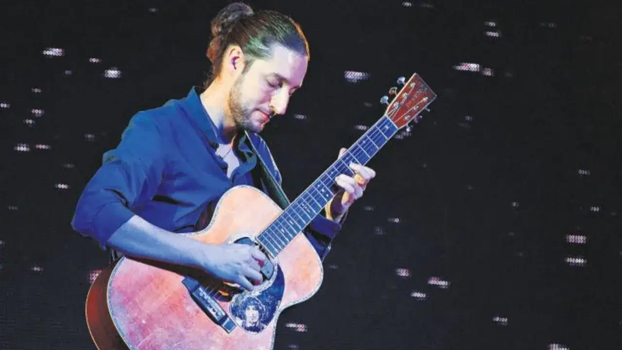 Giovanni Baglioni, con i capelli raccolti, suona la chitarra acustica sul palco, immerso nella musica.