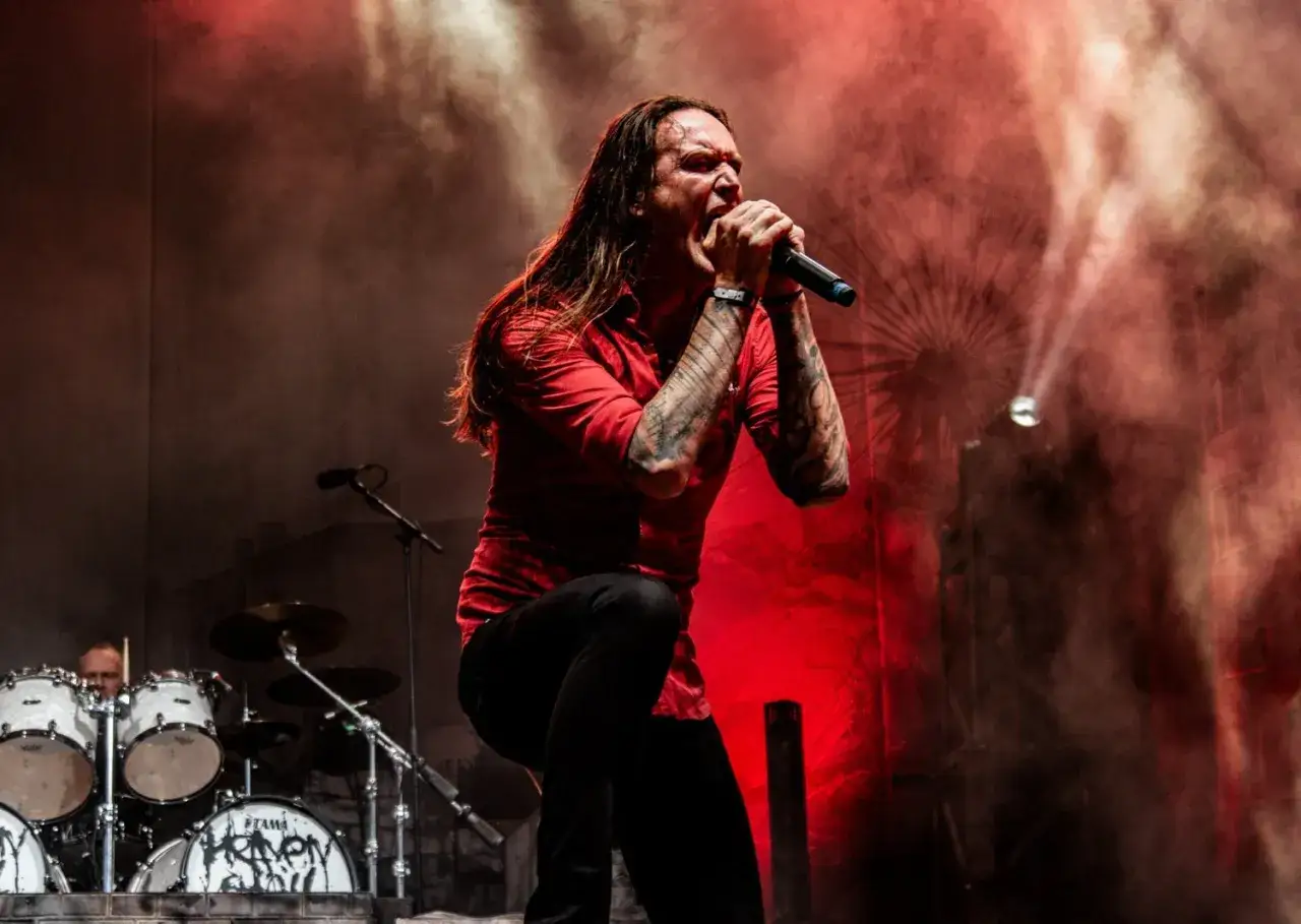 Cantante con lunghi capelli e tatuaggi urla nel microfono, circondato da fumo rosso e luci. Sembra che il heaven shall burn.
