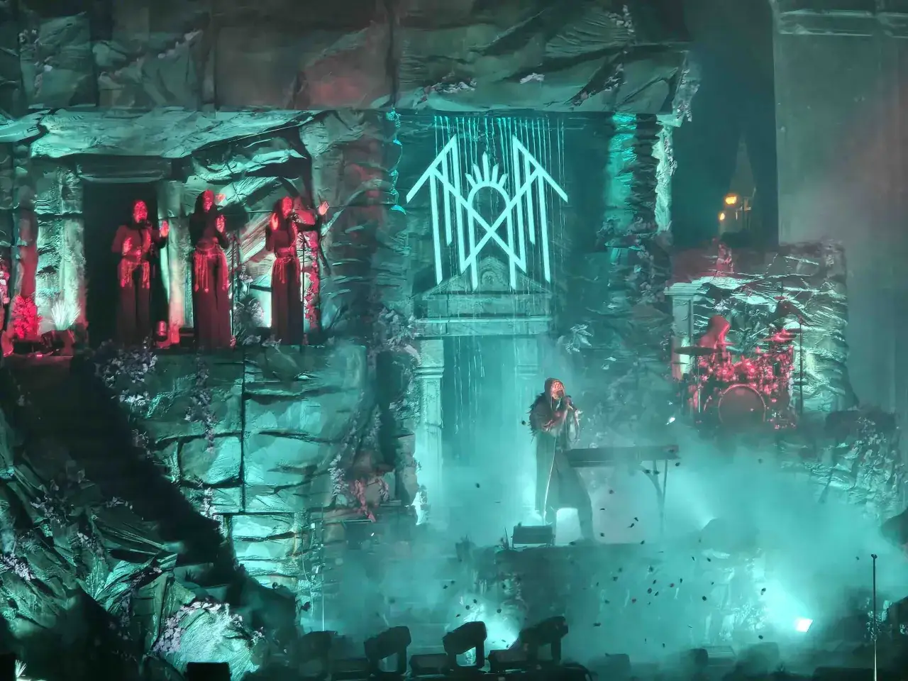 Sul palco, un'atmosfera mistica per il concerto dei Sleep Token. Figure in rosso cantano su piattaforme rocciose, mentre il frontman suona le tastiere tra fumo e luci.