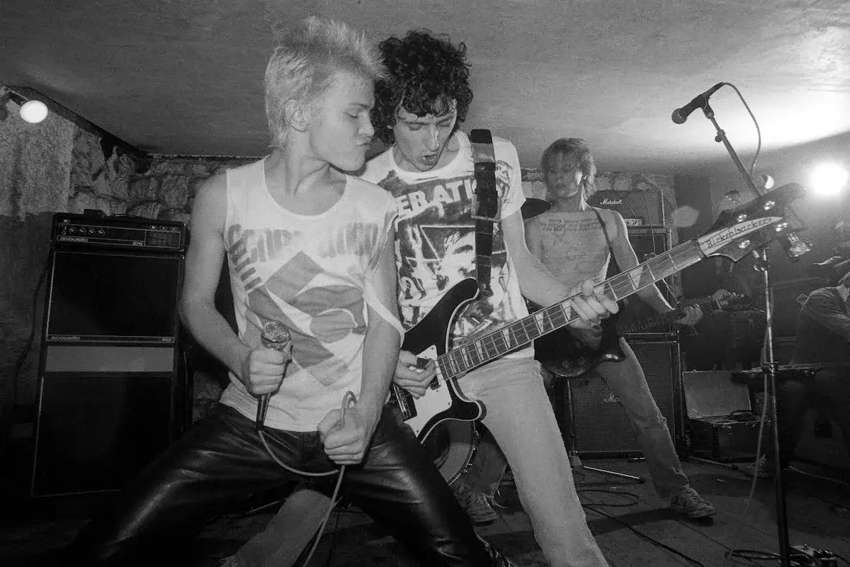 Billy Idol giovane con cresta bionda canta al microfono, mentre un chitarrista suona.