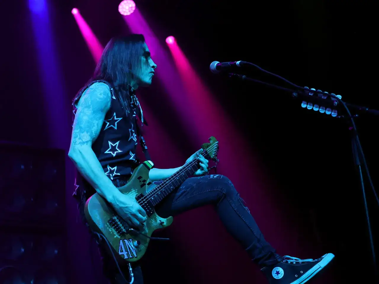 Nuno Bettencourt oggi sul palco, con la sua chitarra verde e un look da rockstar. Luci rosa e blu creano un'atmosfera elettrica.