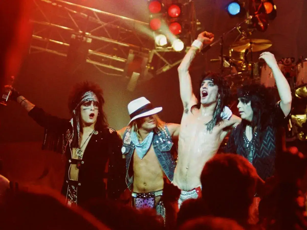 I membri dei M&ouml;tley Cr&uuml;e anni '80 sul palco, con trucco pesante e abiti audaci, celebrano un momento di pura energia rock.