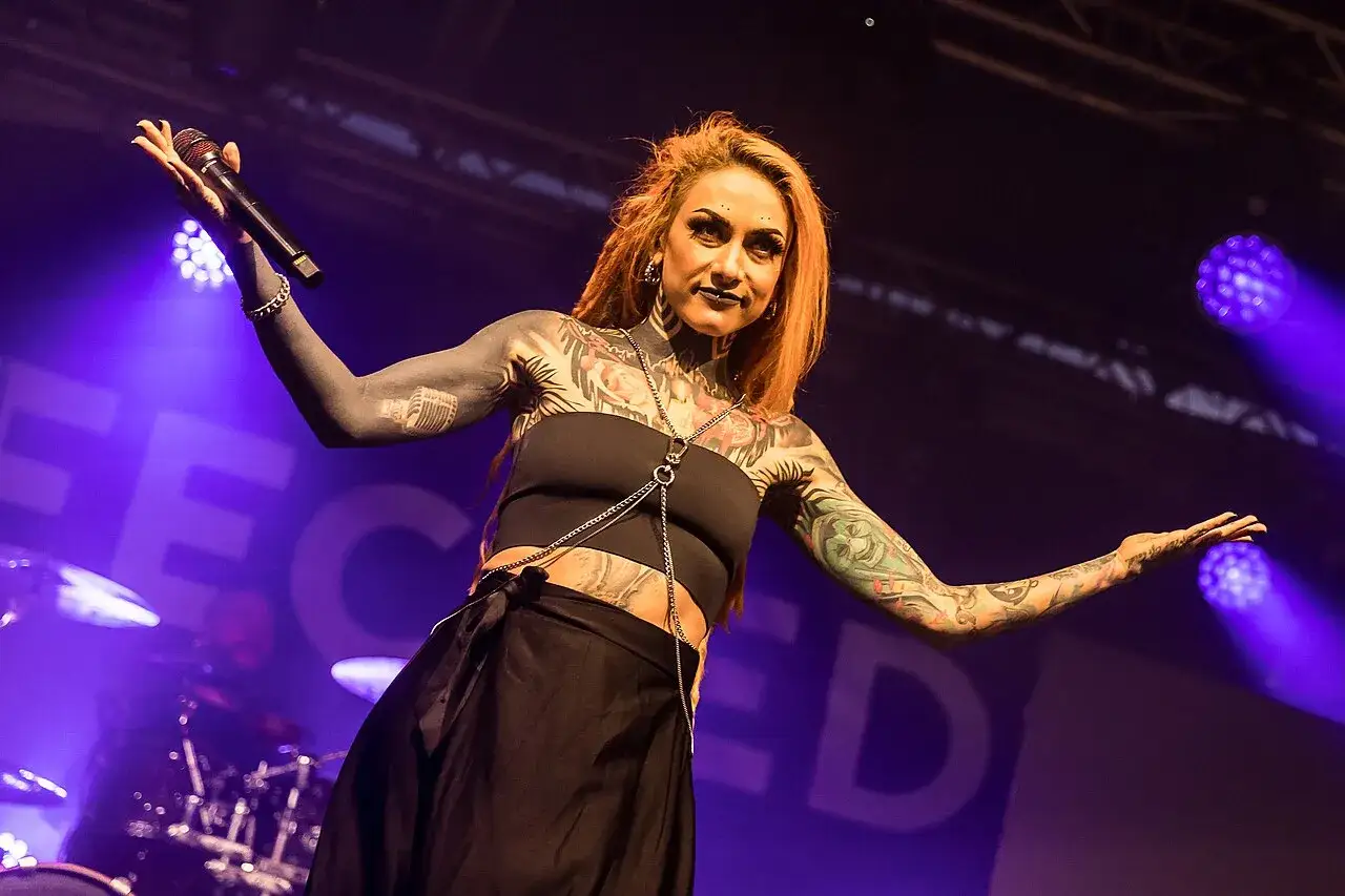 Cantante con tatuaggi su tutto il corpo, microfono in mano, sotto una luce viola che ricorda la pioggia infetta.