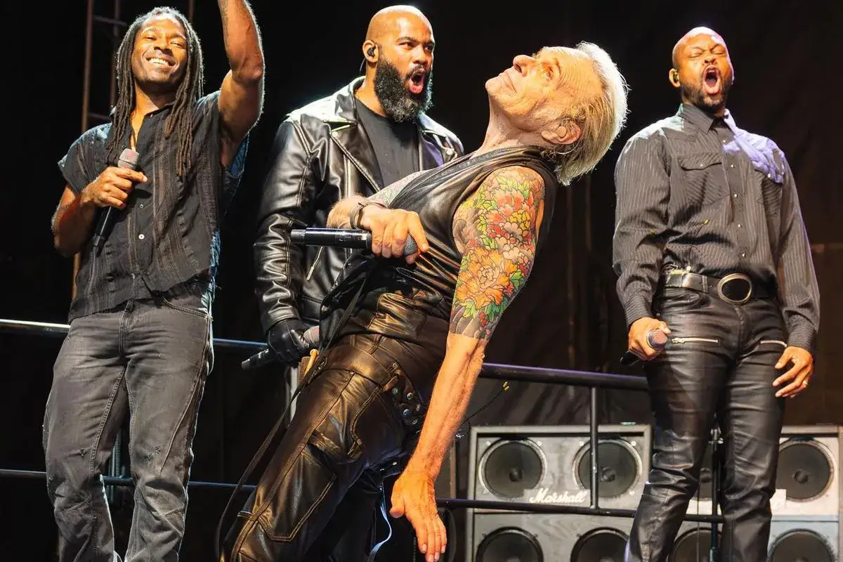 David Lee Roth sul palco, con tatuaggi colorati. Nonostante la sua malattia, continua a esibirsi con energia.