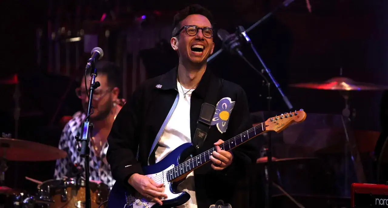 Cory Wong sorride mentre suona la chitarra elettrica blu sul palco, con un batterista sullo sfondo.