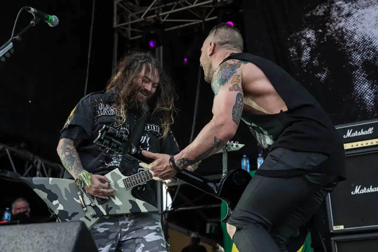 Due chitarristi sul palco, uno con una chitarra mimetica, l'altro con tatuaggi visibili. Sembra che stiano per scontrarsi, un vero
