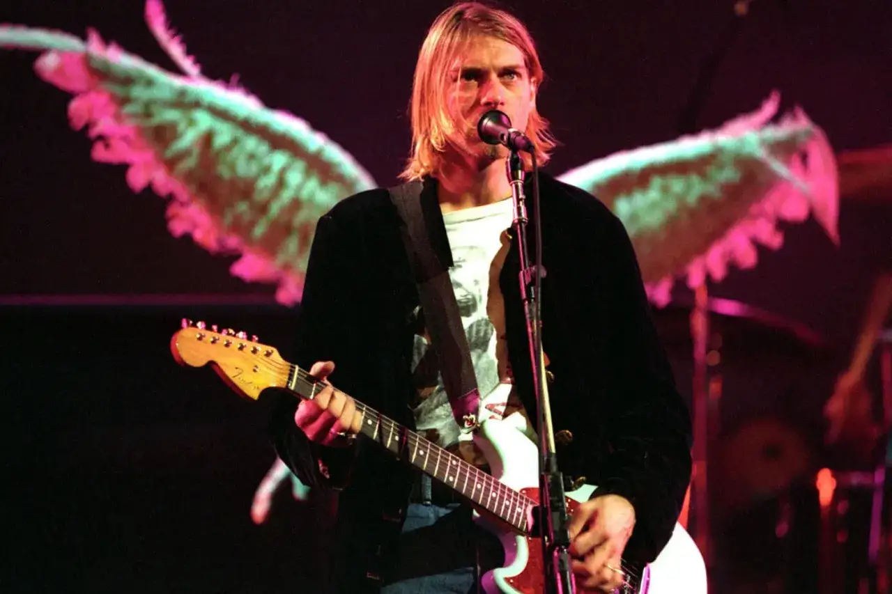 Il chitarrista dei Nirvana, Kurt Cobain, canta sul palco con la sua chitarra. Dietro di lui, un'immagine di ali rosa e verdi.