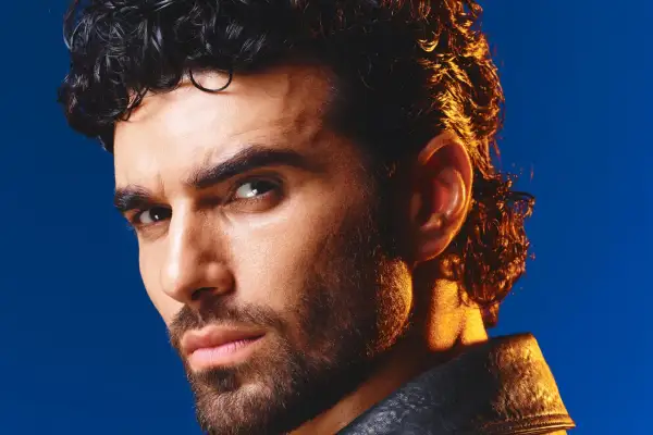 Volto intenso di un uomo con capelli ricci scuri e barba, illuminato da una luce dorata. Ricorda i cantanti di una volta.
