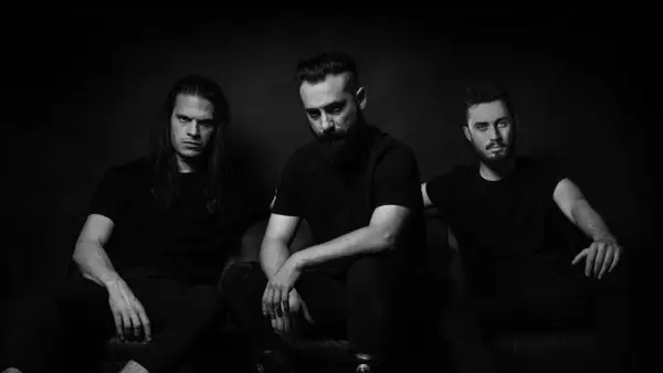 Tre uomini con espressioni intense, vestiti di nero, seduti su un divano. Sembrano il **genus ordinis dei** che si prepara per una performance.