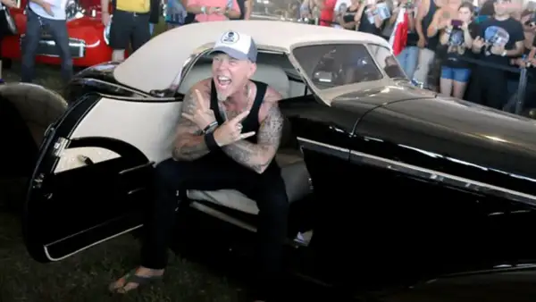 Metallica James Hetfield, con cappellino e canotta, fa il segno delle corna seduto in un'auto d'epoca nera.