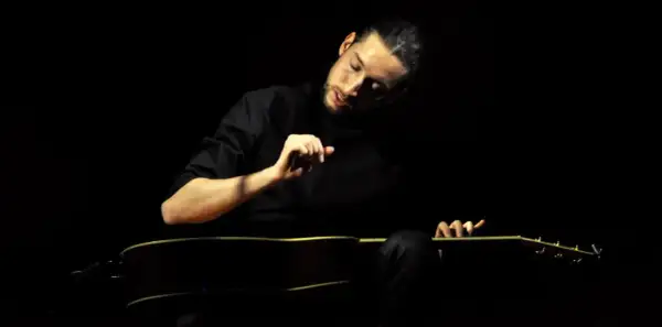 Giovanni Baglioni, con la sua chitarra, crea un'atmosfera intima e intensa. La sua mano destra accarezza le corde, mentre il volto è immerso nella penombra.